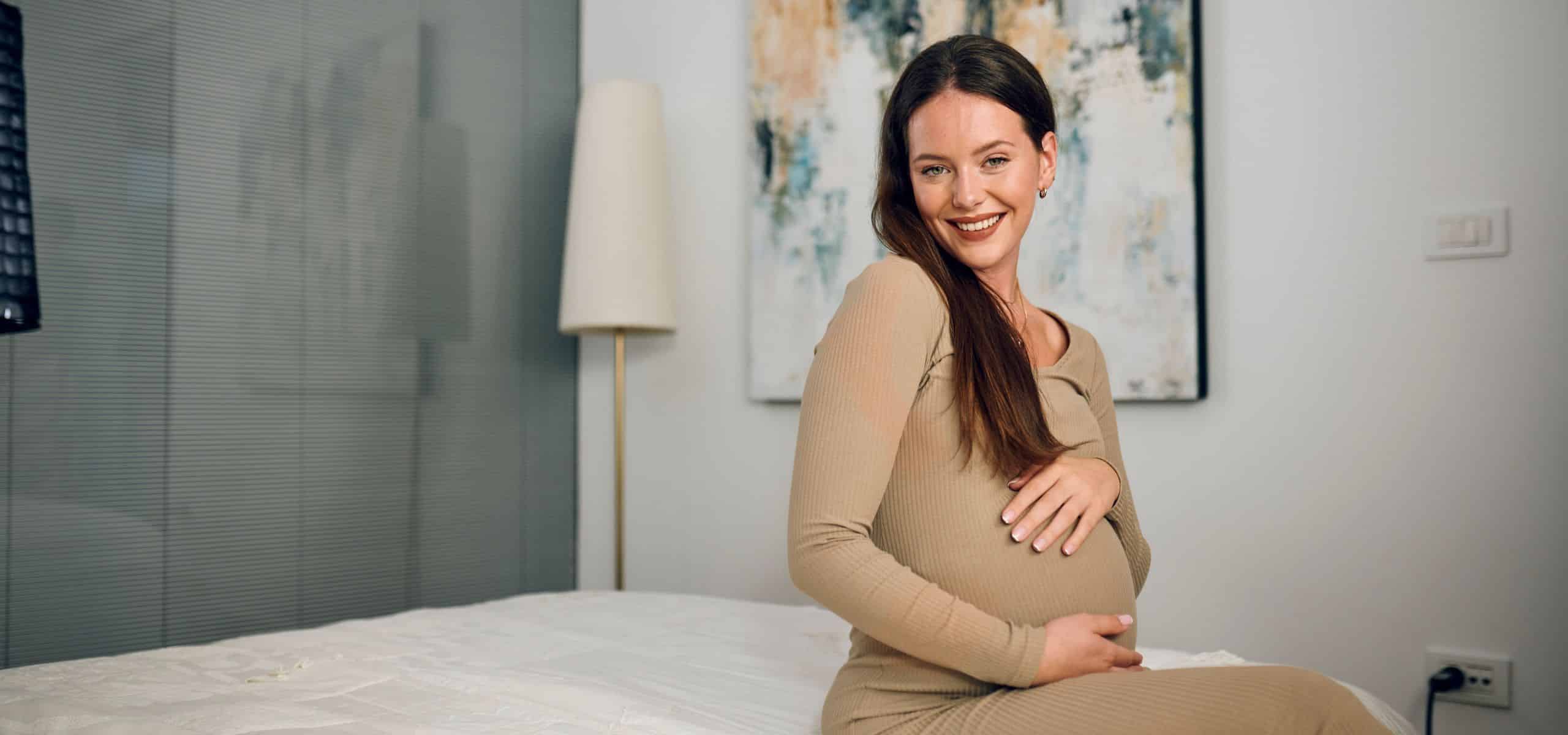 noseča ženska sedi na postelji in se boža po trebuščku